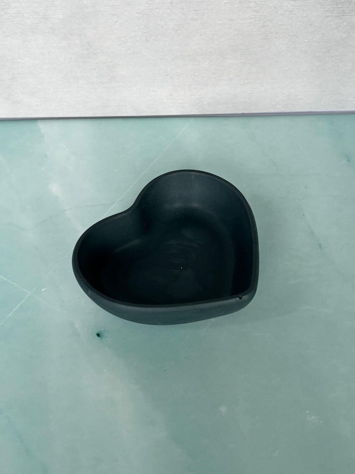 A small matte black heart-shaped trinket bowl or dish placed on a pale green surface with a light gray wall behind it.