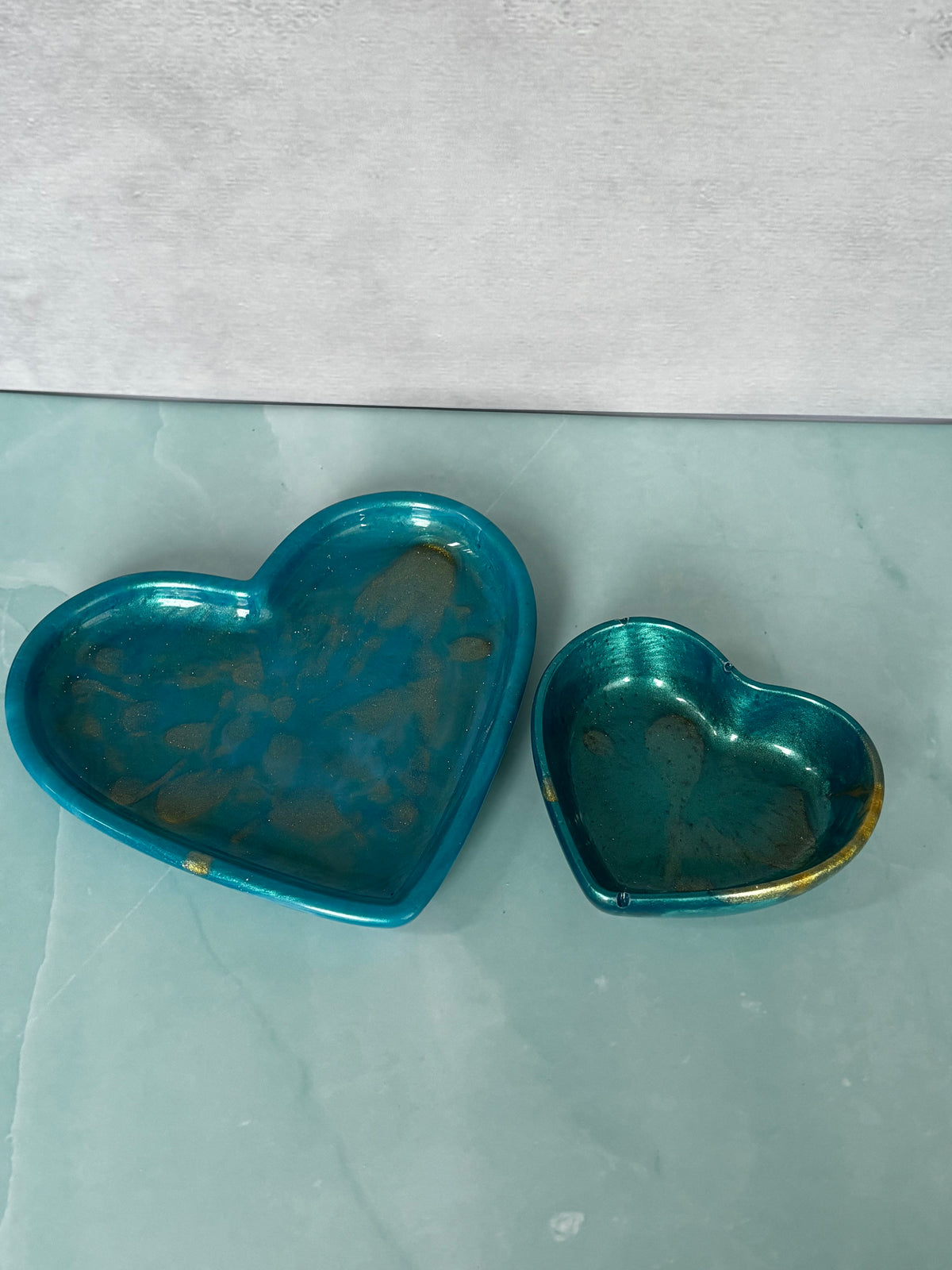Two heart-shaped turquoise glass dishes with gold-toned rims, one larger and one smaller, placed on a light gray marble surface.