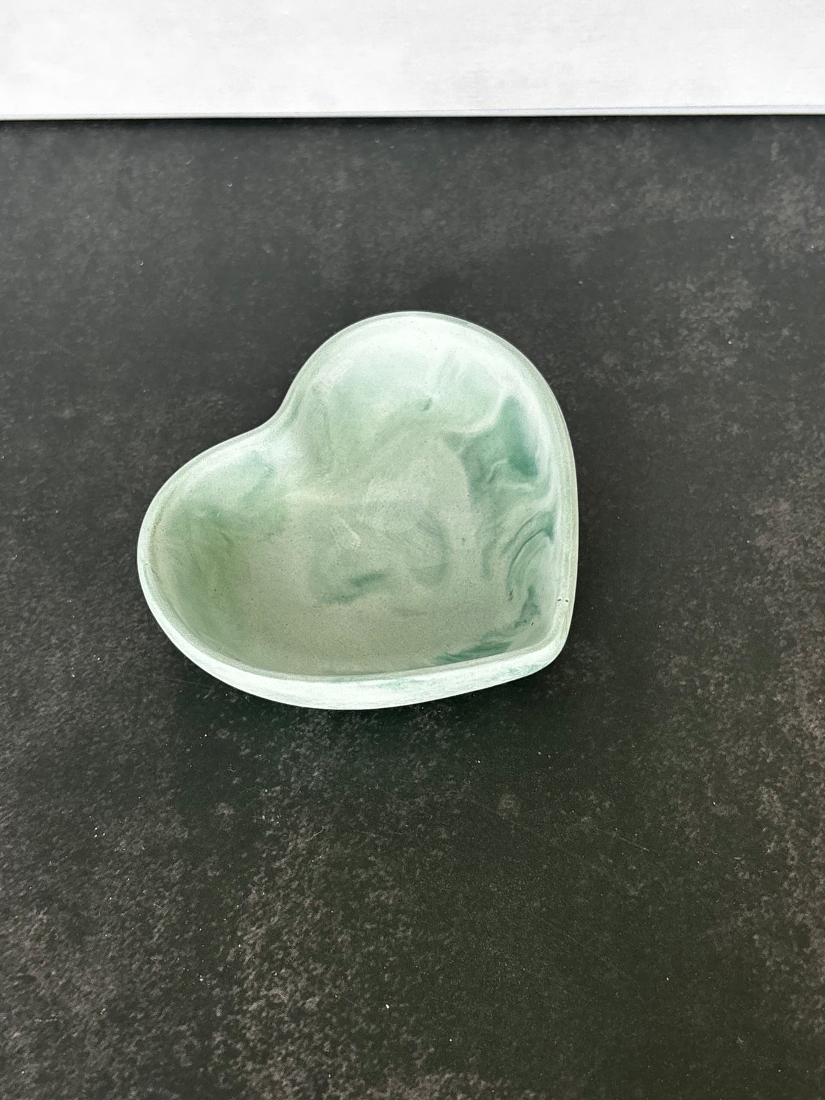A small heart-shaped trinket dish made of pale green marbled hydrostone, shown on a dark textured surface.
