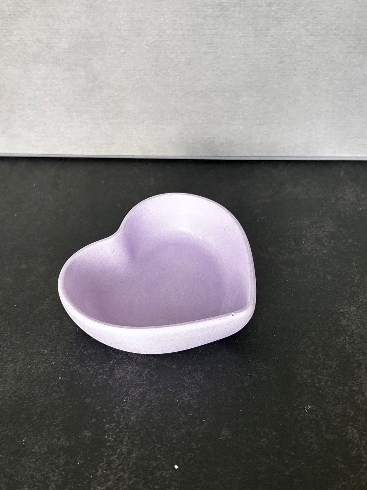 A small heart-shaped trinket bowl or dish in light lavender purple, shown on a dark textured surface with a gray background.