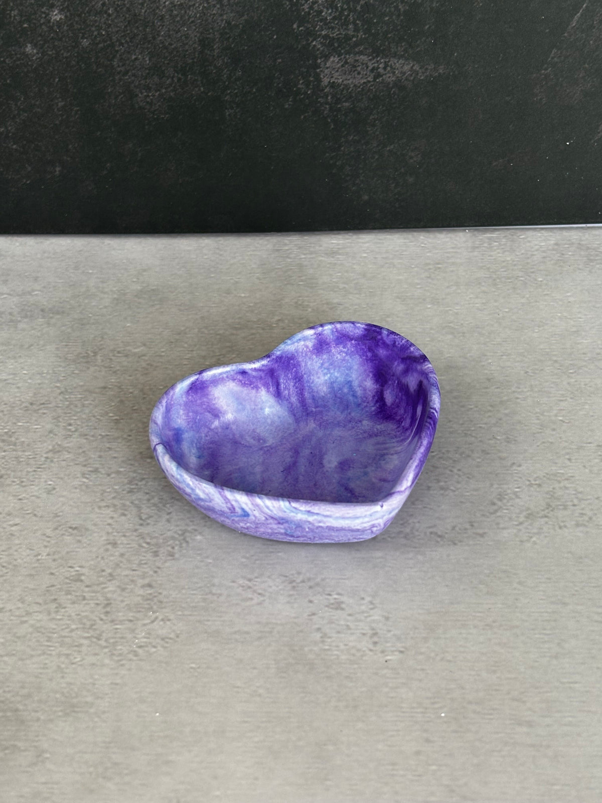 A small heart-shaped trinket bowl or dish with a marbled purple and white pattern, set on a gray surface against a dark gray background.