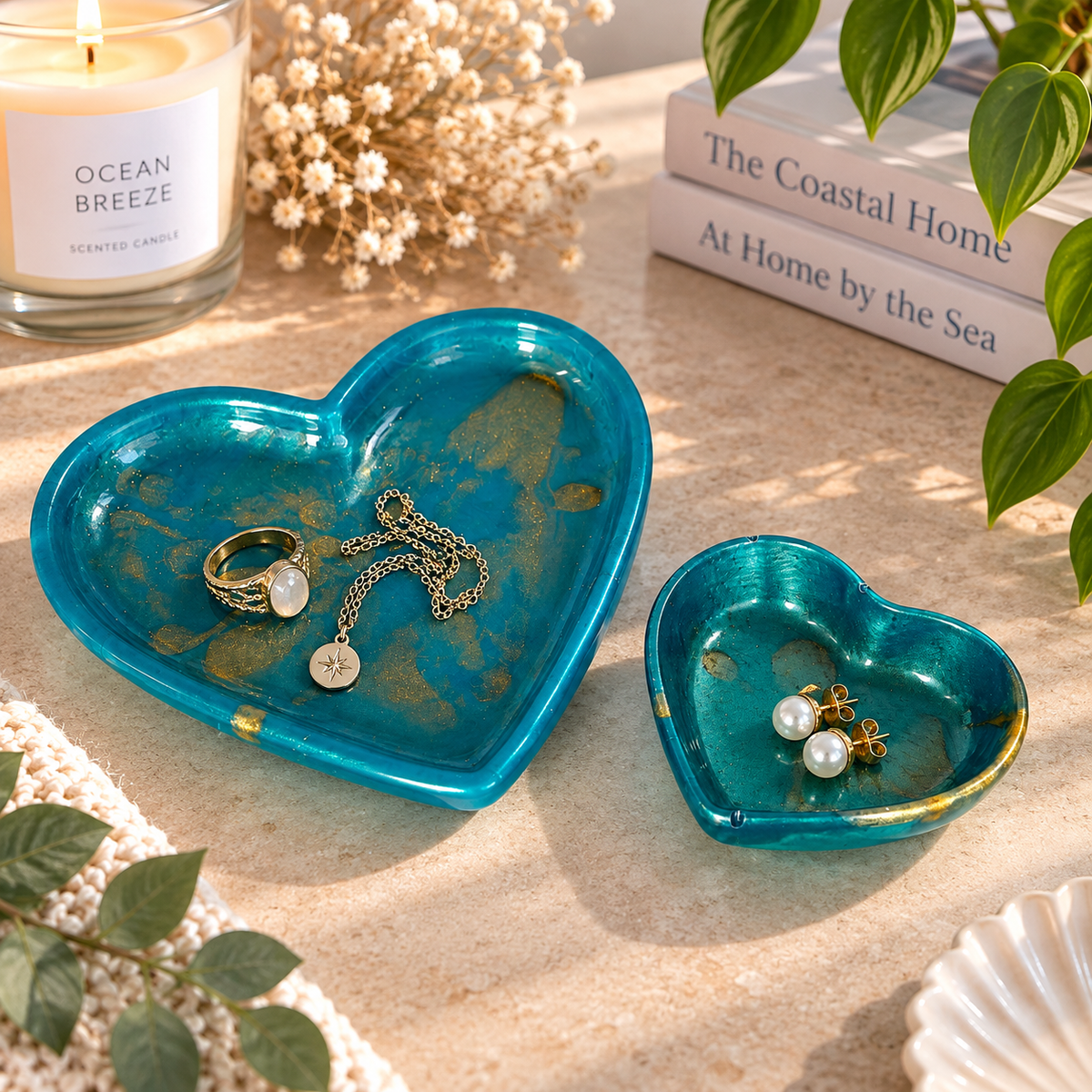 Two turquoise resin heart-shaped trinket trays with gold accents, styled on a beige tabletop. The larger tray holds a ring and necklace, while the smaller tray holds pearl earrings, with a candle, books and greenery in the background.
